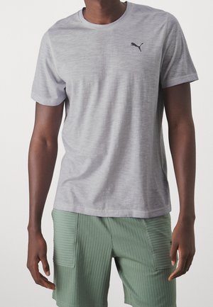 Man draagt een lichtgrijs Puma T-shirt en geribbelde groene korte broek met voorzakken, staand tegen een effen achtergrond.