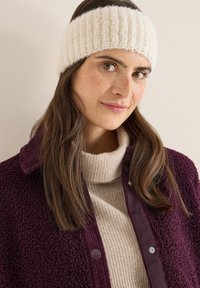 Dziana kremowa opaska na uszy o prążkowanej fakturze; noszona z beżowym swetrem o teksturze i ciemnofioletową kurtką z kołnierzem z sztruksu.