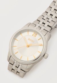 Ασημένιο μεταλλικό ρολόι χειρός με χρυσά σημάδια ωρών και δείκτες, που δείχνει ώρα 11:10:42, με το όνομα της μάρκας "BOSS" σε λευκό καντράν με υφή.