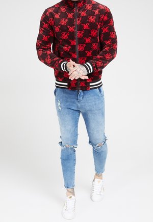 Chaqueta bomber roja y negra con patrón repetido de logo, puños y bajo acanalados. Combinada con vaqueros azules desgastados y zapatillas blancas.