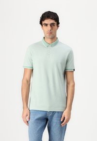 Lys grønn polo skjorte med krage, korte ermer og hvite detaljer. Stoffet ser glatt ut, kombinert med blå denim jeans.