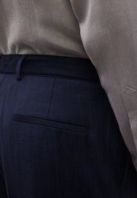 Προσαρμοσμένο σκούρο μπλε παντελόνι με λεπτές λευκές ρίγες και πίσω τσέπη με φιτίλι, φορεμένο με μπεζ πουκάμισο που έχει μαζευτεί μέσα.