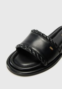 Black slide sandal with a thick braided strap, smooth leather texture, rounded toe, and subtle gold logo detail on the side.