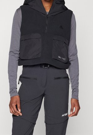 Osoba ubrana w czarną bezrękawnikową kamizelkę z kapturem i przednimi kieszeniami na szarej koszuli z długim rękawem oraz czarne spodnie zapinane na zamek z logo na udzie.