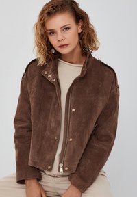 Bruin suède militaire stijl jack met een ritssluiting aan de voorkant, gouden hardware en schouderepauletten. Gladde textuur, cropped ontwerp en relaxte pasvorm.