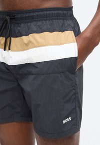 Κοντινό πλάνο ενός προσώπου που φορά μαύρο μαγιό σορτς με μπεζ και λευκές οριζόντιες ρίγες και λογότυπο "BOSS" στο πόδι, με το χέρι στην τσέπη.