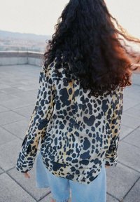 Camicia in stampa leopardo in tessuto morbido, con vestibilità ampia e maniche lunghe. I colori includono beige, nero e toni terrosi attenuati.