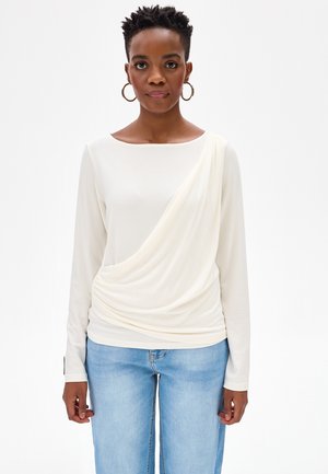 Vrouw met kort krullend haar, gouden oorringen in de vorm van hoepels, een wit langarmig gedrapeerd shirt en lichtblauwe spijkerbroek, staand tegen een witte achtergrond.