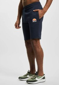 Pantalones cortos azul marino con cintura elástica y bolsillos laterales, que presentan un logo rojo, naranja y blanco en el muslo izquierdo.