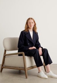 Manteau bleu marine croisé porté sur un haut blanc, associé à un pantalon noir. Assis sur une chaise rembourrée de couleur claire avec un cadre en bois.