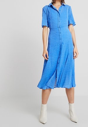 Kobieta ubrana w niebieską sukienkę midi na guziki z krótkim rękawem i małym wzorem w kropki oraz białe botki, stojąca na jednolitym tle.