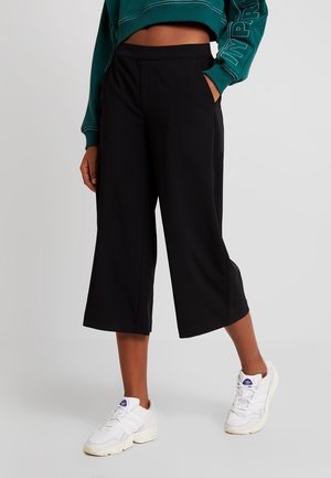 Person, die schwarze, weit geschnittene, kurze Hose, ein türkisfarbenes kurzes Sweatshirt mit Schrift auf dem Ärmel und weiße Sneakers trägt, steht vor einem schlichten Hintergrund.