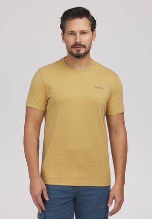 Mężczyzna z krótkimi ciemnymi włosami i brodą, ubrany w jednolity żółty podkoszulek i niebieskie spodnie, stoi na tle jasnoszarego tła.