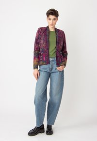 Veste à motifs colorés avec une base sombre, comportant des motifs complexes et deux poches avant, associée à une chemise verte et un pantalon large.