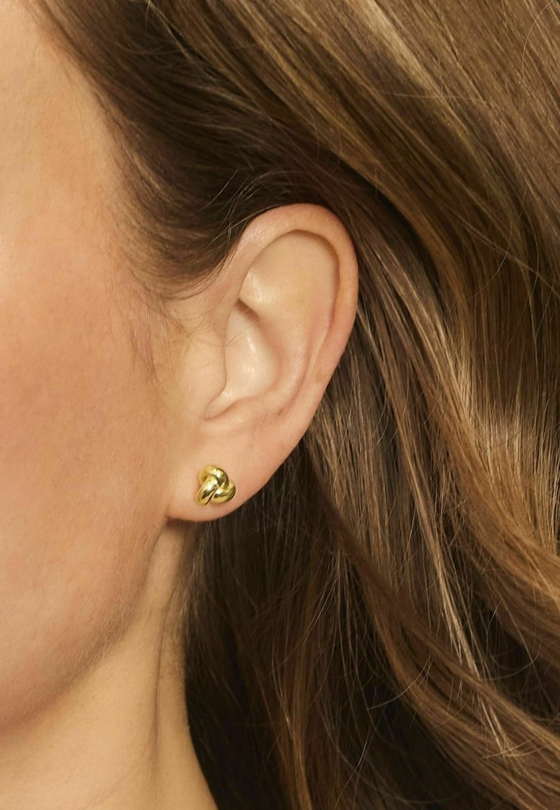 Orecchini a perno in oro con un design intrecciato, caratterizzati da una superficie liscia e riflettente e una finitura lucida. Indossati all'orecchio.