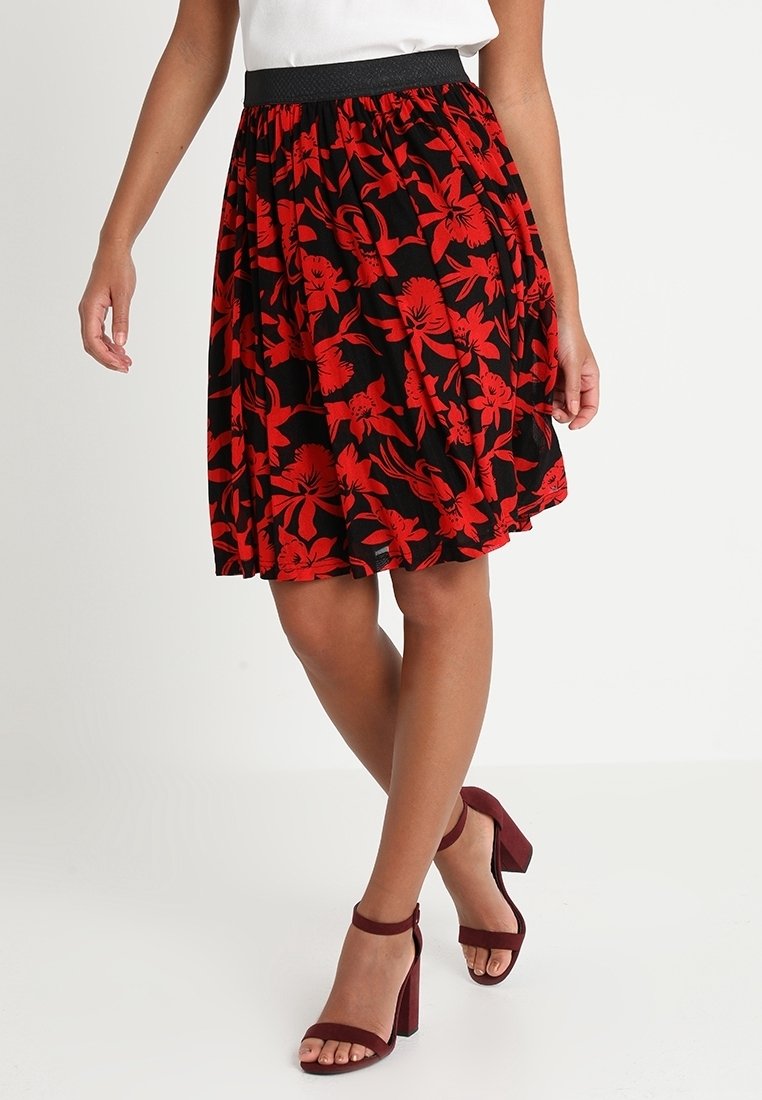 Jupe florale mi-longue avec une base noire et de belles fleurs rouges. Taille élastique. Associée à des sandales à talons bordeaux.