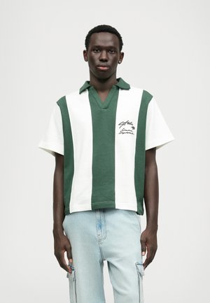 Polo vert et blanc à rayures, à manches courtes et col en V, avec un détail de texte brodé sur le côté gauche.