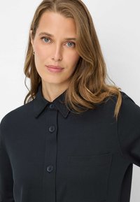 Sort skjorte med knapper, krave, lomme på venstre bryst og glat stoftekstur. Modellen bærer den med løst hår.