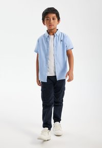 Chemise à manches courtes bleu clair avec des boutons, T-shirt blanc en dessous, pantalon cargo bleu marine foncé et baskets blanches avec des accents gris.
