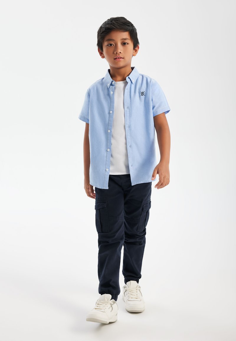 Chemise à manches courtes bleu clair avec des boutons, T-shirt blanc en dessous, pantalon cargo bleu marine foncé et baskets blanches avec des accents gris.