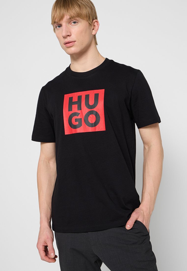 Camiseta de algodón negra con un cuadrado rojo que tiene el texto "HUGO" en negrita negro. Mangas cortas, escote circular, ajuste relajado.
