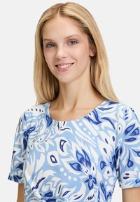 Květinové šaty s krátkým rukávem ve světle modré látce, s bílým a tmavě modrým vzorem; kulatý výstřih a hladká textura.
