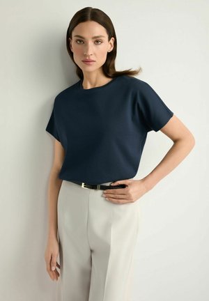 Kobieta o prostych brązowych włosach, ubrana w granatową bluzkę z krótkim rękawem wsuniętą w jasnobeżowe spodnie z czarnym paskiem, stojąca na tle jasnej ściany.