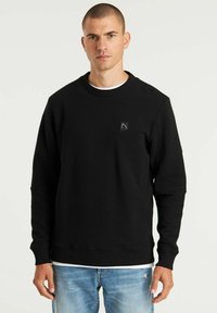 Schwarzer gerippter Sweatshirt mit rundem Halsausschnitt, langen Ärmeln und einem kleinen Logo-Patch auf der Brust, kombiniert mit hellblauen, ausgewaschenen Jeans.