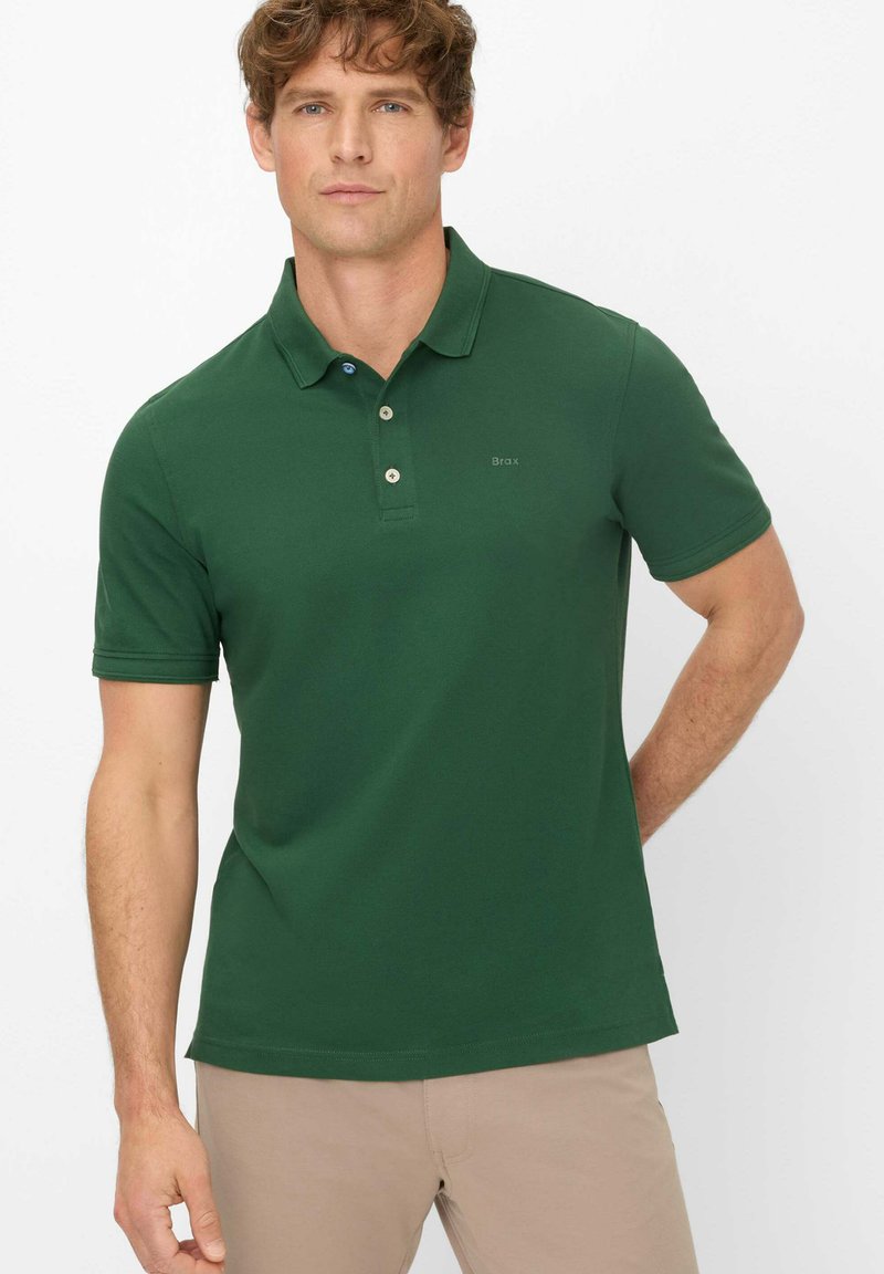 Dunkelgrünes Poloshirt mit kurzen Ärmeln, dreiknöpfigem Kragen, dezentem Branding und glatter Textur, kombiniert mit hellbeigen Hosen.