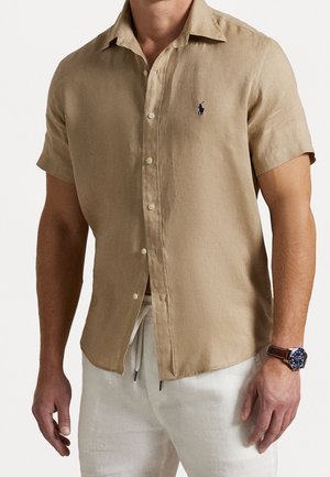 Homme portant une chemise beige à manches courtes avec un petit logo sur la poitrine, un pantalon blanc et une montre-bracelet avec un bracelet en cuir.