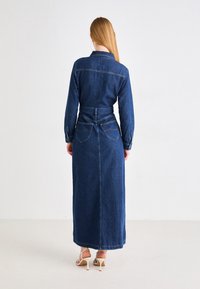 Vestido maxi de mezclilla en azul oscuro, con mangas largas, cintura ceñida y bolsillos en la parte trasera. Textura suave con costuras visibles.