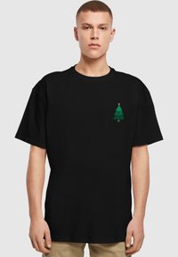 Sort T-shirt med korte ærmer, der har et lille grønt juletræsgrafik på venstre brystområde, lavet af bomuldsstof.