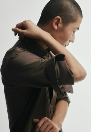 Jauns cilvēks ar īsiem matiem regulē viņa tumši brūnas, uzrullētu piedurkņu krekla apkakli, stāvot sānis pret vienveidīgu fonu.