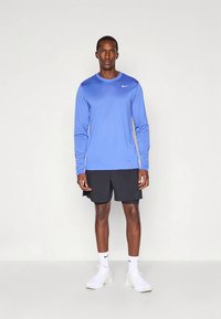 Langermet blå treningsskjorte med rund hals og liten hvit Nike-logo, kombinert med svarte shorts og hvite joggesko.
