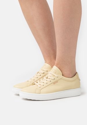 Beige low-top leather sneakers with white soles and laces worn on bare feet against a white background.