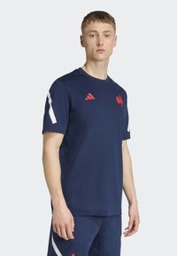 Mörkblå sporttröja med kort ärm och röda accenter, inklusive en logotyp och ränder på ärmarna, tillverkad av ett mjukt, andningsbart material.