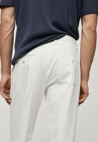 Białe, teksturowane spodnie z tylnymi kieszeniami i jasnymi guzikami, zestawione z ciemną bluzką z krótkim rękawem. Widok z tyłu.