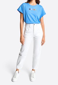 Ljusblå bomullströja med korta ärmar och broderade smileys, kombinerad med vita jeans och vita sneakers.