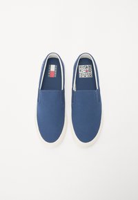 Zapatillas de lona azul tipo slip-on, con suela de goma blanca, textura suave y un código QR en el lado interno para información del producto.