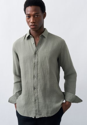 Joven con una camisa de manga larga color verde claro abotonada, con las manos en los bolsillos, de pie frente a un fondo claro y liso.