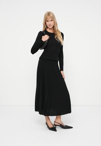 Γυναίκα με μακριά ξανθά μαλλιά, φορώντας μαύρη μπλούζα με μακριά μανίκια, μαύρη πλισέ μέση φούστα, μαύρες ψηλές τακούνια slingback, κρατώντας μια μαύρη τσάντα ώμου.