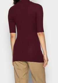 Žena oblečená v přiléhavém vínovém svetr s krátkým rukávem a béžových kalhotách, na obrázku ze zadu proti jednoduchému pozadí.