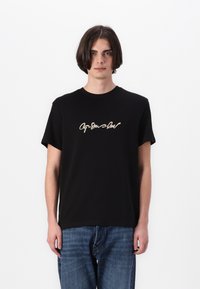 T-shirt en coton noir à manches courtes, avec un design de texte en cursive crème sur la poitrine. Porté avec un jean en denim bleu.