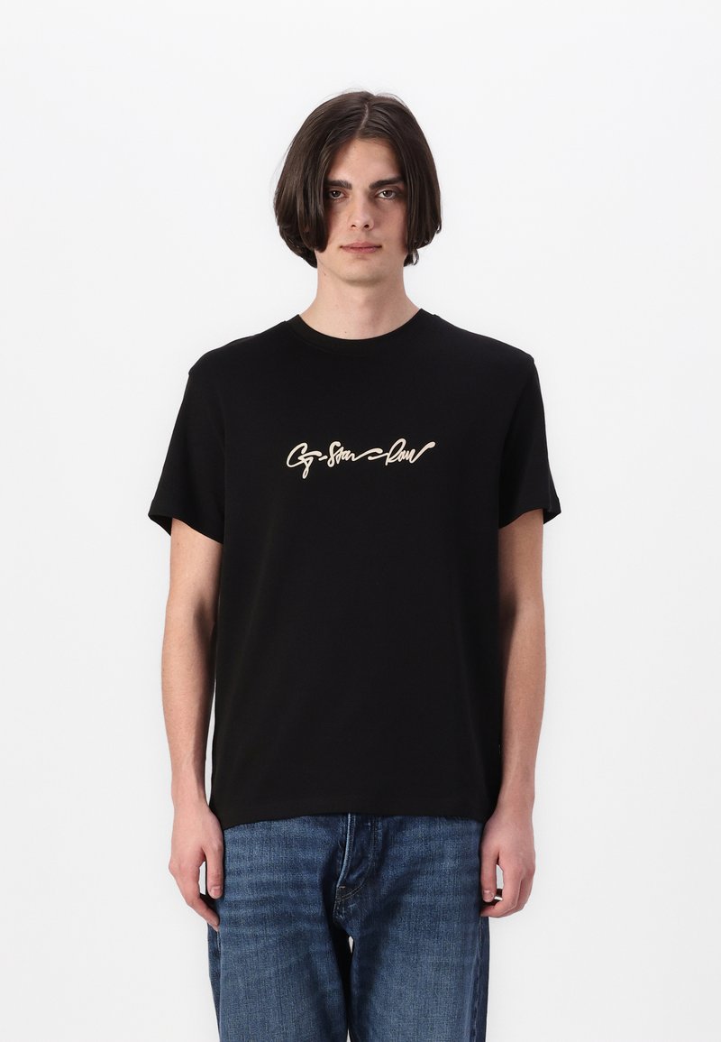 T-shirt en coton noir à manches courtes, avec un design de texte en cursive crème sur la poitrine. Porté avec un jean en denim bleu.