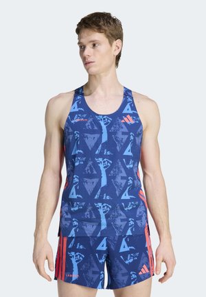 Νεαρός άνδρας που φοράει αθλητική φανέλα με μπλε σχέδια και ταιριαστά σορτς με κοραλί ρίγες, στέκεται και κοιτάζει αριστερά πάνω σε απλό φωτεινό φόντο.