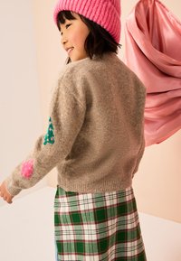 Maglione color beige in misto lana con collo rotondo, caratterizzato da toppe verdi e rosa sulle maniche, abbinato a una gonna a quadri verde. Berretto rosa in maglia.