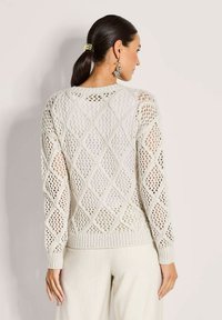 Frau mit dunklem Pferdeschwanz, die einen cremefarbenen offenen Strickpullover und helle Hosen trägt, von hinten vor einem schlichten hellen Hintergrund gezeigt.