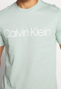 Camiseta de cuello redondo en verde menta hecha de tela suave, que presenta un gran logo blanco de "Calvin Klein" en el pecho. Manga corta.