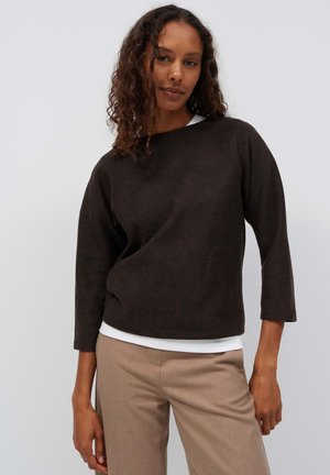Donna con capelli ricci che indossa una maglia a maniche lunghe marrone scuro sopra una camicia bianca e pantaloni beige, in piedi davanti a uno sfondo chiaro e uniforme.