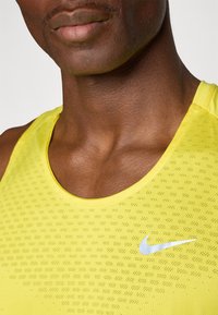 Gul atletisk tanktopp med et teksturert mønster, rund hals, og en hvit Nike-logo på venstre side. Mykt stoff, ermeløs design.